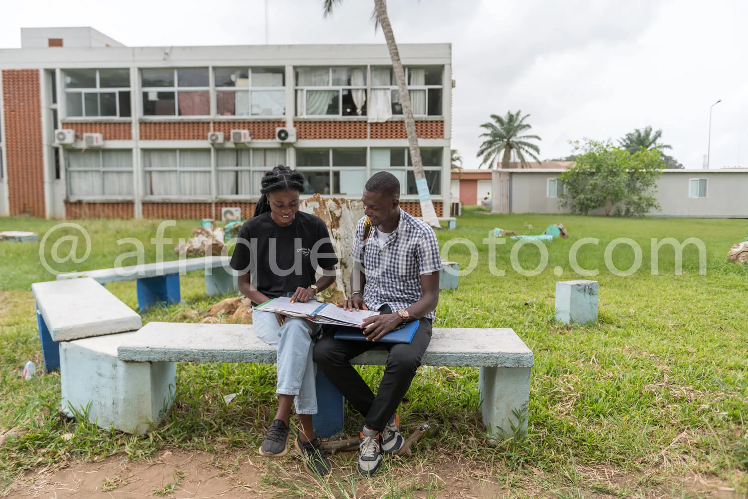 Africa photo — Deux personnes assises sur un banc en béton, souriantes, étudiant un cahier ouvert ensemble dans un parc avec des arbres et un bâtiment