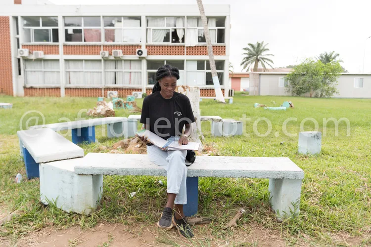 Africa photo — Une personne assise sur un banc en béton dans un espace vert, tenant un carnet ouvert et écrivant avec un stylo, devant un immeuble à é