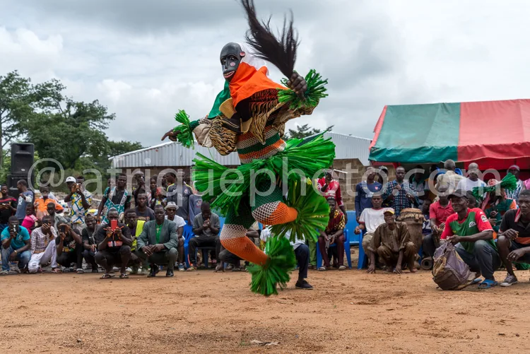 Africa photo — Un danseur traditionnel vêtu d’un costume aux couleurs vives, vert, orange et jaune, exécute une danse dynamique devant un public assis