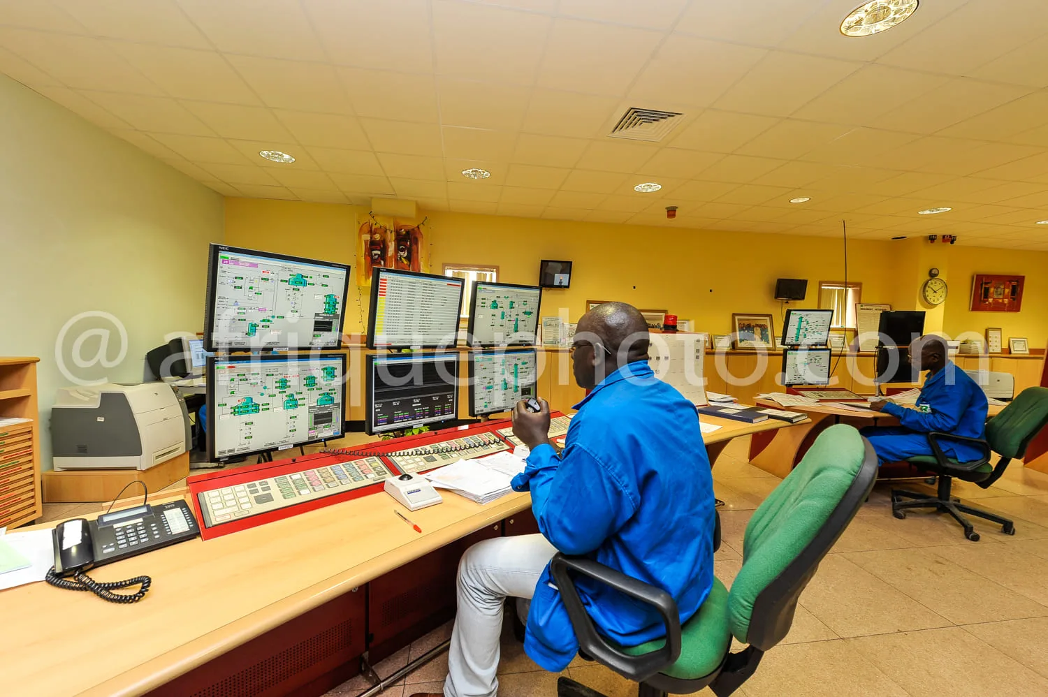 Africa photo — Deux opérateurs en combinaison bleue travaillent dans une salle de contrôle industrielle équipée de plusieurs écrans affichant des sché