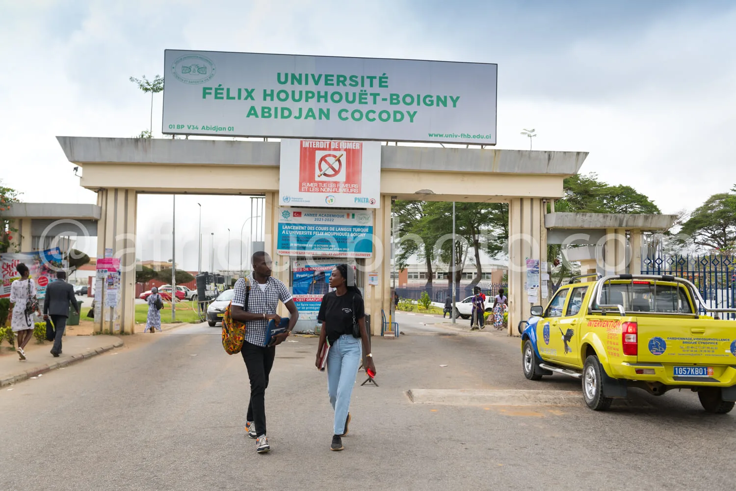 Africa photo — Vue de l’entrée principale de l’université Félix Houphouët-Boigny à Abidjan Cocody, avec deux étudiants marchant vers la sortie, un pan