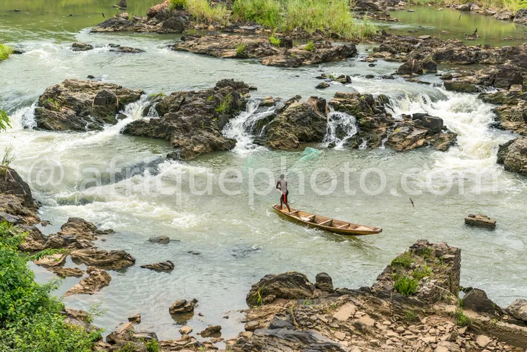 Africa photo — Un pagayeur en canoë traditionnel navigue sur une rivière aux eaux vives, entourée de rochers moussus et de végétation luxuriante, dans