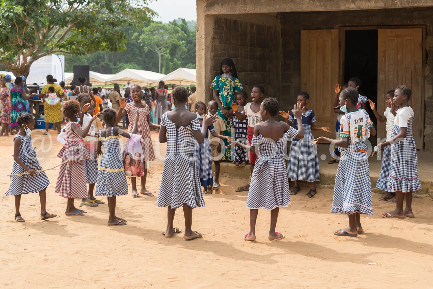Africa photo — Un groupe d’enfants vêtus d’uniformes scolaires à carreaux bleus et blancs, dansant en cercle sur une terre battue devant une école en