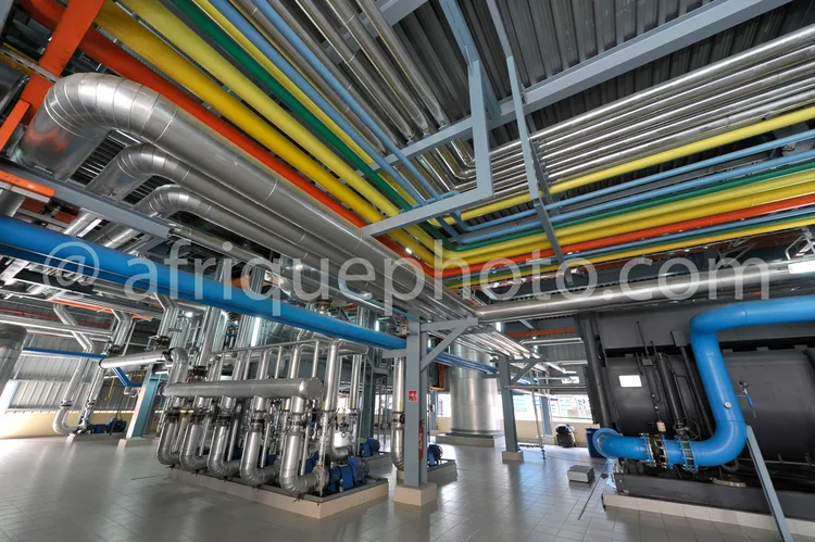 Africa photo — Intérieur d'une usine industrielle montrant un réseau complexe de tuyaux colorés (bleu, gris, jaune) et gaines techniques suspendues au