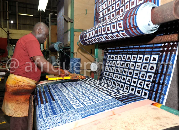 Africa photo — Un artisan en tenue de travail, vêtu d’un t-shirt rouge et d’un tablier, manipule une machine à tisser pour créer un tissu aux motifs g