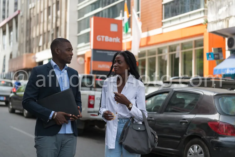 Africa photo — Un homme en costume bleu et chemise blanche, tenant un dossier, discute avec une femme en veste blanche et jean, devant une enseigne GT