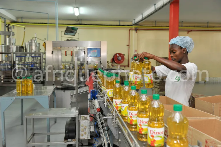 Africa photo — Une employée en tenue blanche et coiffe professionnelle vérifie des bouteilles d'huile végétale Dinor sur une chaîne de production indu