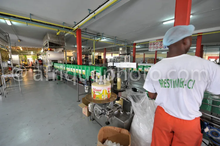 Africa photo — Un salarié en tenue de travail blanche et orange, coiffe blanche, se tient près d'une ligne de production industrielle. On aperçoit des