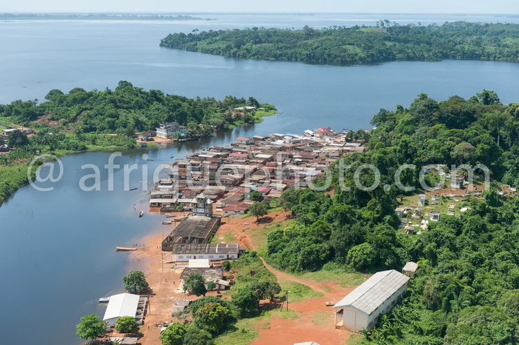 Africa photo — Vue aérienne d’un village situé au bord d’un grand lac, entouré de forêts denses. On aperçoit des maisons en bois et en tôle, des bâtim