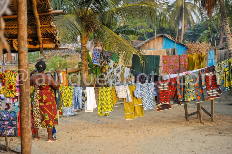 Africa photo — Une femme vêtue d’une robe traditionnelle africaine à motifs colorés se tient près d’un étalage de tissus wax suspendus, aux couleurs v