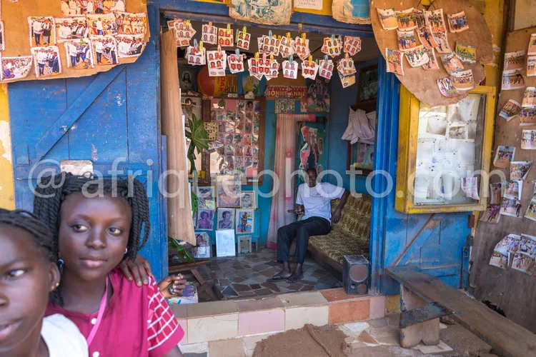 Africa photo — Deux enfants se tiennent devant une boutique aux murs bleus et jaunes, couverts de photos encadrées et de petits objets décoratifs. À l
