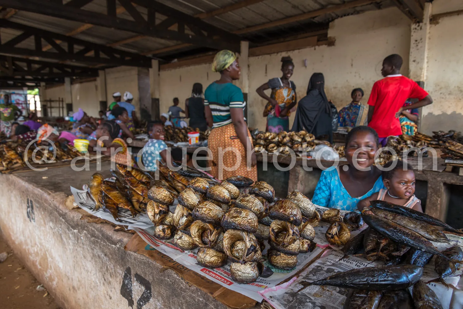 Africa photo — Une scène animée d’un marché artisanal où des vendeuses souriantes présentent des boules alimentaires dorées et fumées, disposées sur d