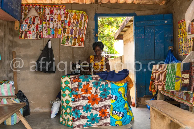 Africa photo — Une femme assise dans un atelier artisanal, entourée de tissus wax aux motifs floraux et géométriques colorés, avec des peintures mural