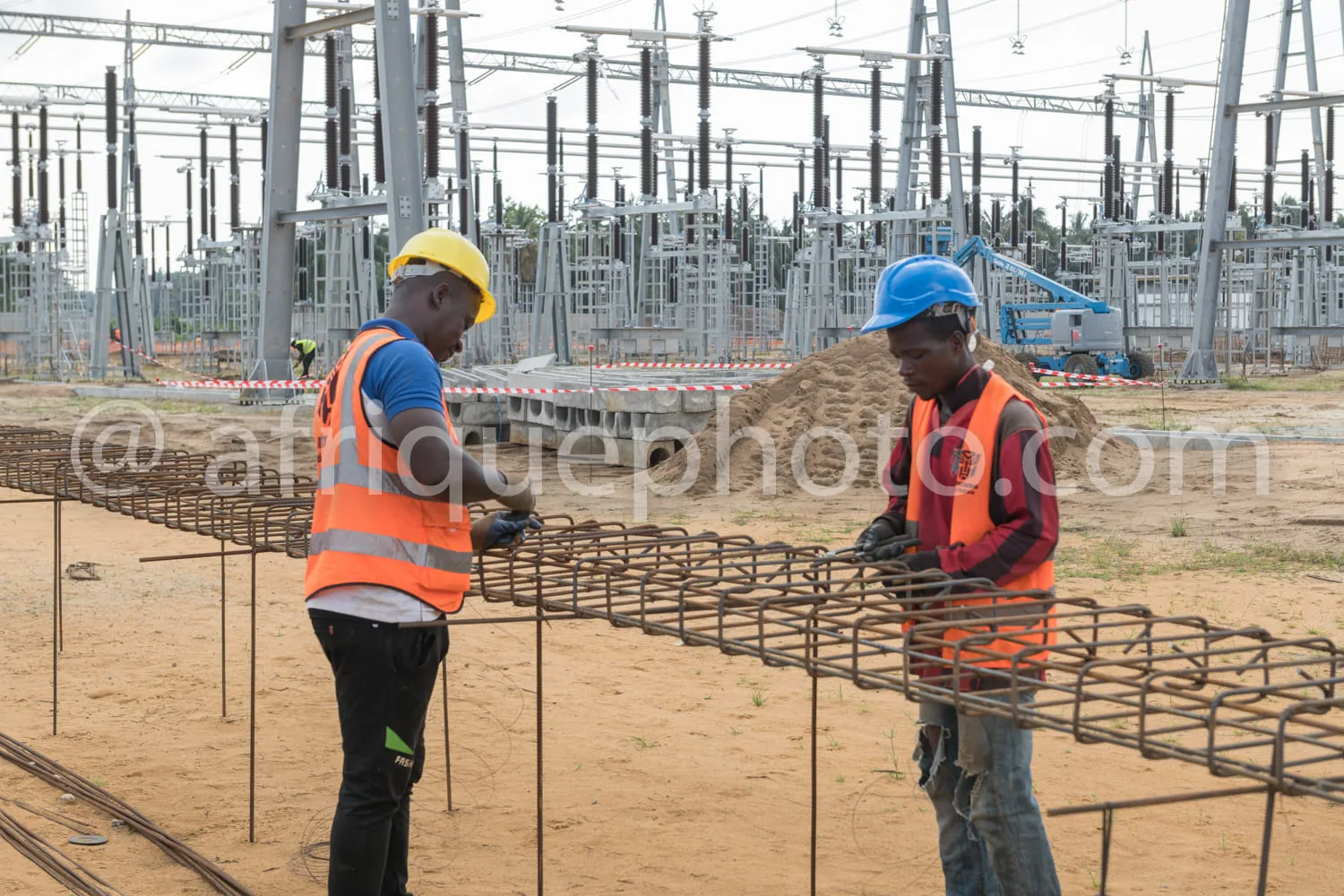 Africa photo — Deux ouvriers en sécurité sur un chantier de construction, portant casques et gilets orange, assemblant des armatures en acier sur un s