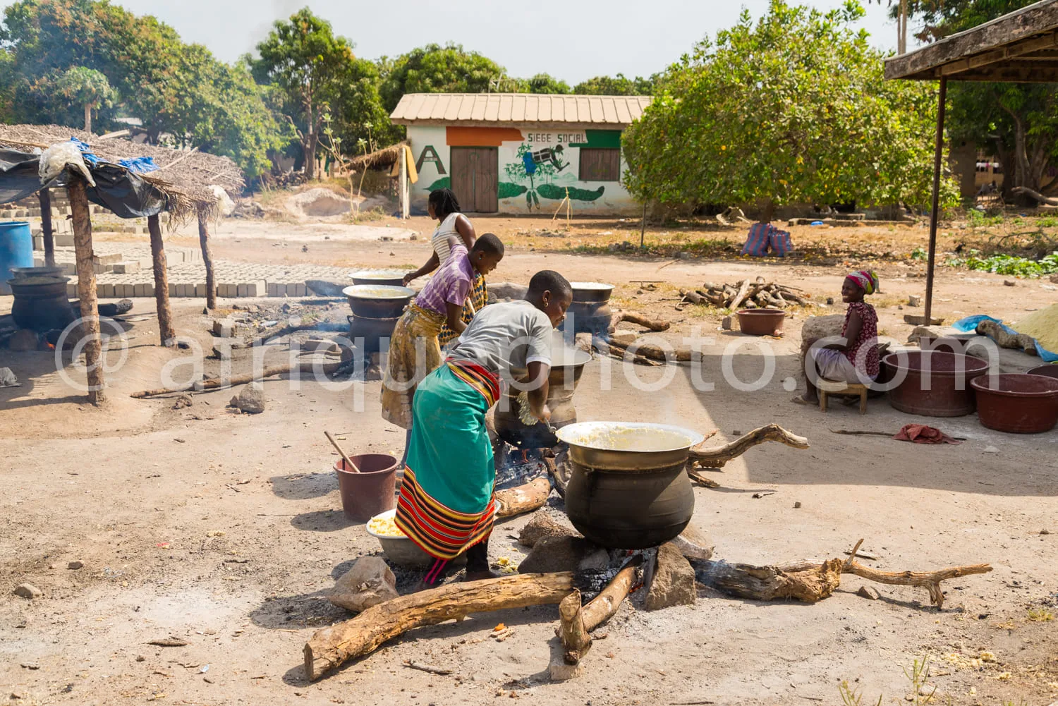 Africa photo — Trois femmes en tenue traditionnelle préparent un repas en plein air autour d’un feu de bois. Elles manipulent des grands pots en métal