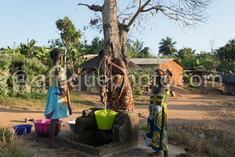 Africa photo — Trois femmes se tiennent autour d’un point d’eau naturel. Une personne actionne une pompe en bois pour remplir un seau vert, tandis que