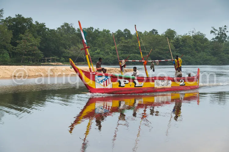 Africa photo — Une pirogue colorée aux teintes rouge, jaune et blanche, décorée de motifs traditionnels, avance sur un plan d’eau calme. À bord, cinq