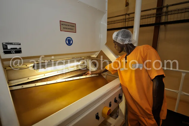 Africa photo — Un technicien vêtu d'une combinaison orange et d'un bonnet jetable inspecte une machine industrielle ouverte, probablement utilisée pou