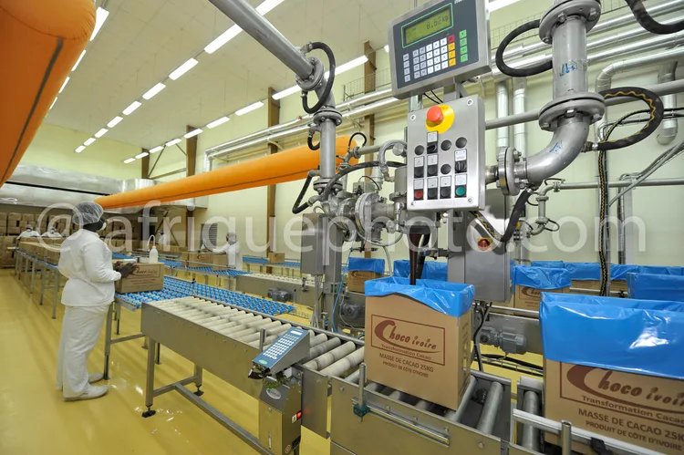 Africa photo — Une ouvrière en combinaison blanche surveille une ligne de production industrielle où des tablettes de chocolat Choco Ivoir sont emball