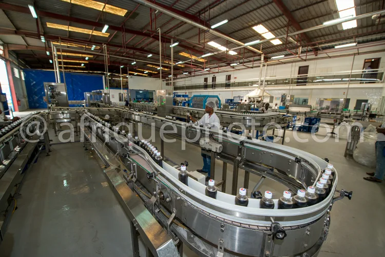 Africa photo — Un opérateur en combinaison blanche surveille une chaîne de production industrielle où des bouteilles en verre sont remplies et bouchée