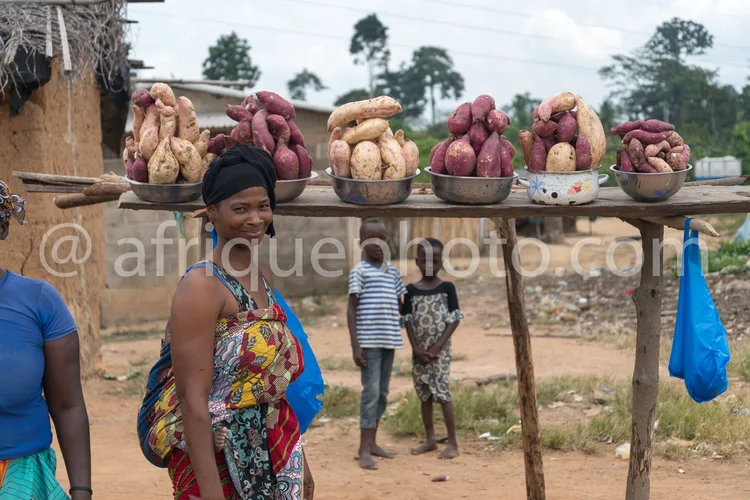 Africa photo — Une femme souriante, vêtue d’un tissu coloré et d’un foulard, porte un plateau rempli de patates douces de différentes couleurs (blanch