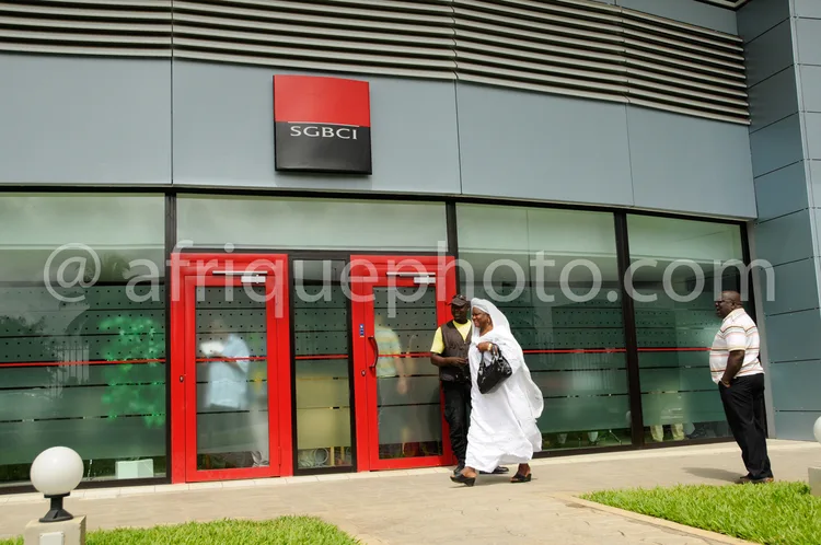 Africa photo — Une femme vêtue d’une tenue traditionnelle blanche entre dans une agence bancaire SGBCI aux portes rouges, accompagnée d’un homme en co