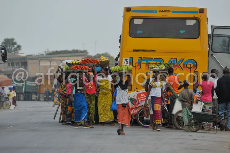 Africa photo — Quatre femmes en tenues colorées transportent des paniers de fruits frais sur la tête et un vélo chargé de marchandises, près d’un cami