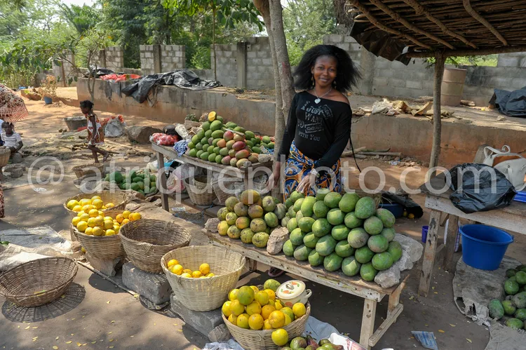 Africa photo — Une femme souriante se tient derrière un étalage de fruits frais, avec des paniers remplis de mangues vertes, de citrons jaunes et de m