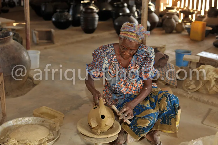Africa photo — Une femme souriante, vêtue d’un tissu coloré et d’un foulard en tête, est assise sur le sol en terre battue. Elle façonne soigneusement