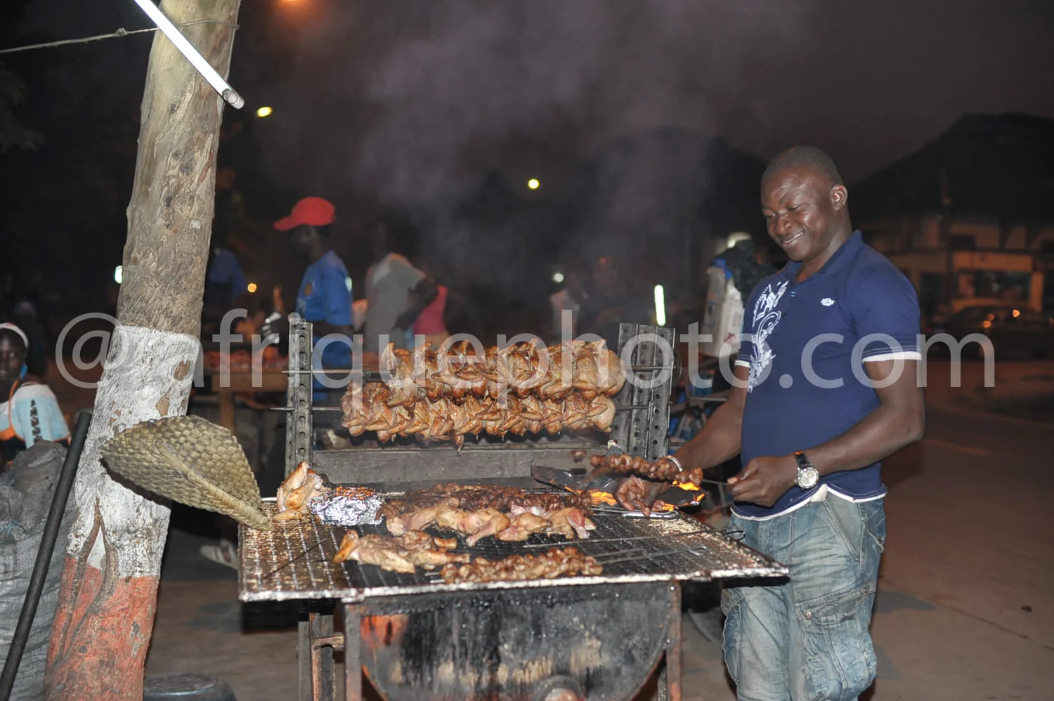 Africa photo — Un homme souriant prépare des brochettes de viande grillées sur un grand barbecue en plein air, entouré de fumée et de lumière nocturne