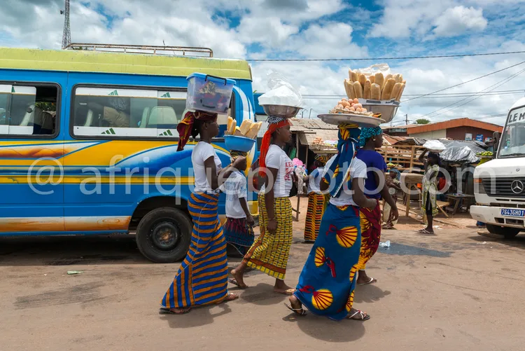 Africa photo — Trois femmes en tenues traditionnelles colorées marchent sur une route poussiéreuse. Elles transportent des paniers de bananes et d'aut