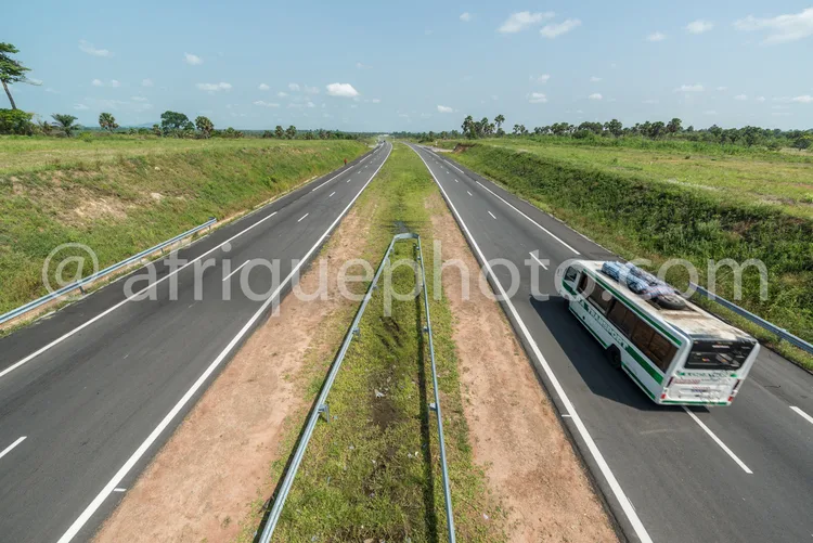 Africa photo — Une route goudronnée large et droite s’étend à l’infini sous un ciel bleu, bordée de végétation verte. Un bus de transport circule sur