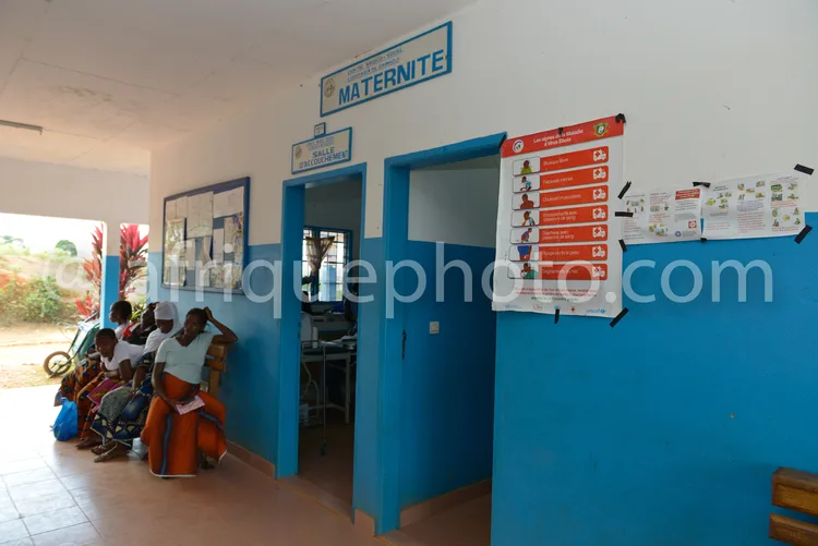 Africa photo — Une salle d’attente de maternité aux murs bleus, avec des femmes assises sur des bancs en plastique orange, une porte bleue marquée « M