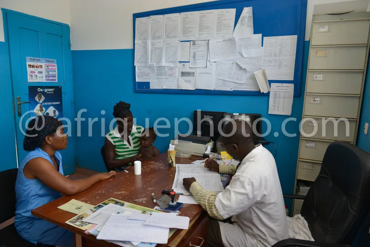 Africa photo — Une salle de consultation médicale bleue où trois personnes sont assises autour d’une table en bois. Une professionnelle de santé en bl