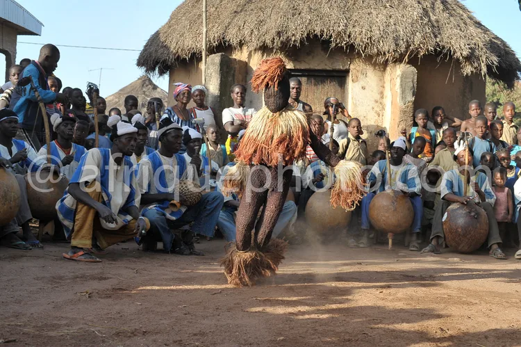 Africa photo — Un danseur traditionnel vêtu d’un costume richement décoré avec des plumes et des motifs colorés exécute une danse devant une foule ass