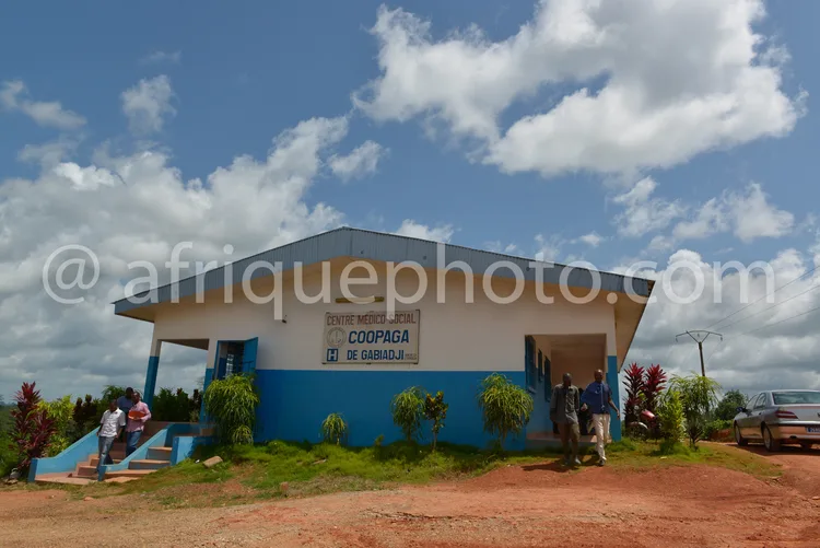 Africa photo — Bâtiment bleu et blanc du centre médical social Coopaga situé à Gabiadji, sous un ciel dégagé avec des nuages. Deux personnes montent l