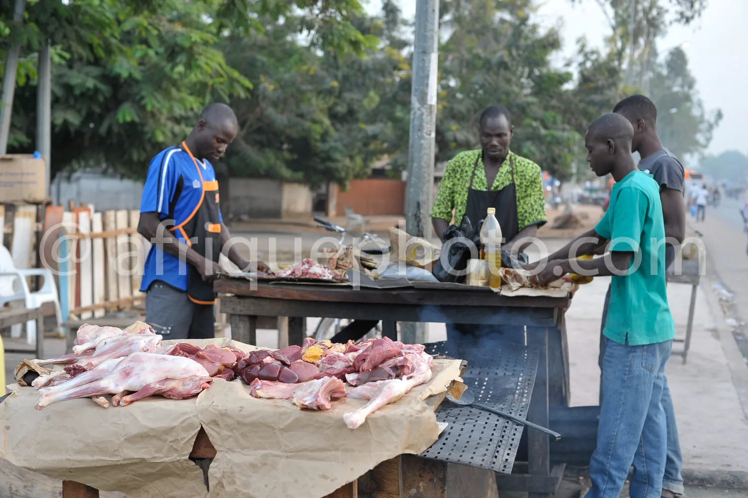 Africa photo — Un étal de boucherie en extérieur où un vendeur, vêtu d’un tablier, prépare de la viande fraîche sur une table grillagée. Trois clients