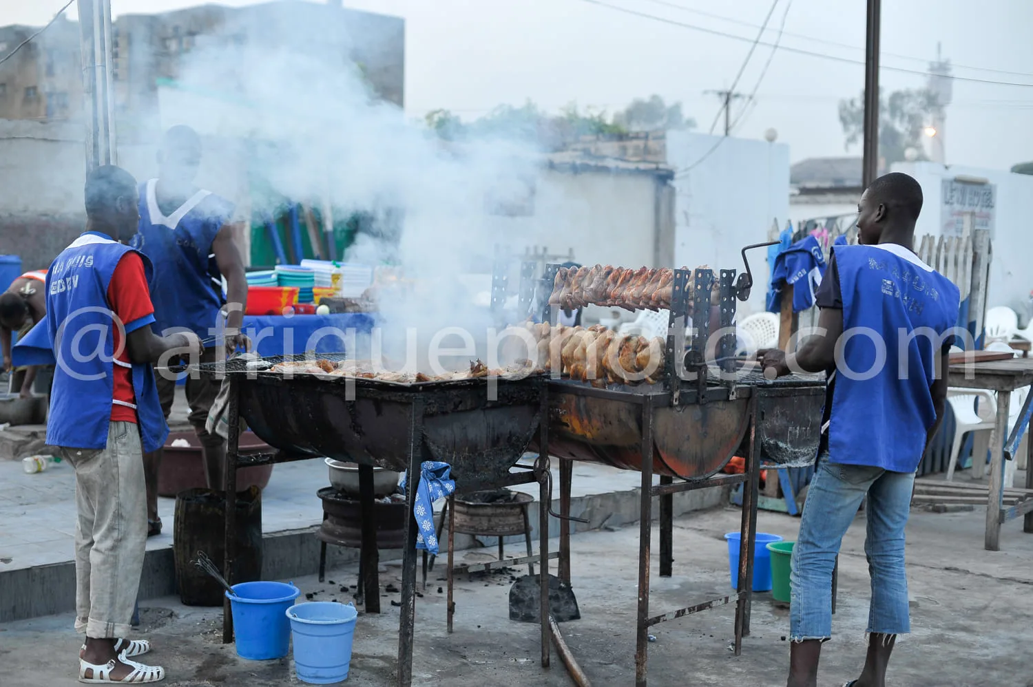 Africa photo — Deux hommes en tenues bleues et rouges manipulent des brochettes de viande grillée sur un grand grill extérieur fumant, entourés de sea