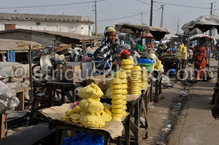 Africa photo — Une femme souriante derrière un étal de marché rempli de tissus colorés, coiffée d’un foulard, entourée de produits artisanaux comme de