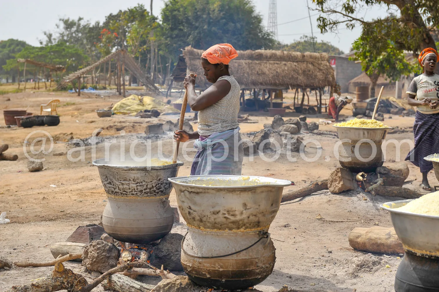 Africa photo — Une femme en tenue traditionnelle remue une grande marmite en terre cuite posée sur un feu de bois, entourée d'autres ustensiles et mar