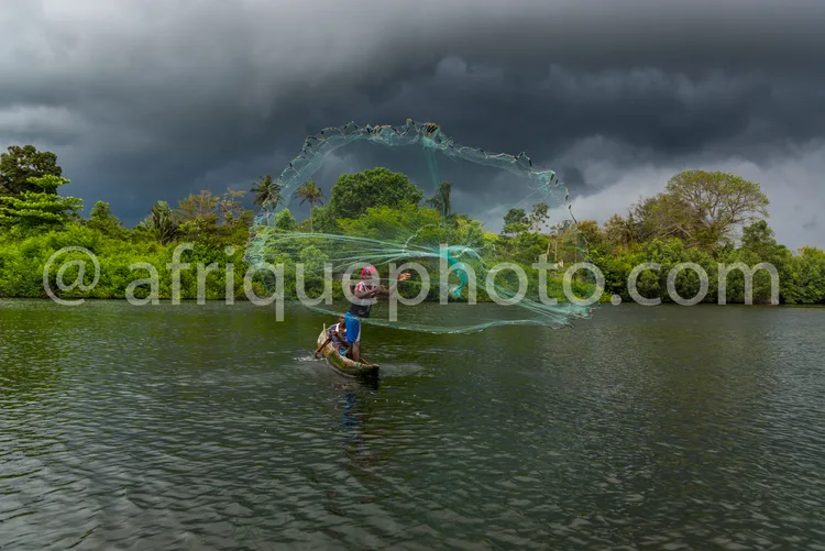 Africa photo — Un pêcheur, vêtu d'un short bleu et d'un chapeau rouge, se tient dans une eau trouble et lance un grand filet circulaire autour de lui,