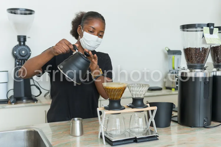 Africa photo — Une barista portant un masque facial verse soigneusement de l’eau chaude dans un filtre à café posé sur un porte-filtre en métal, entou
