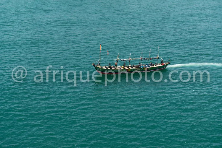 Africa photo — Un bateau en bois traditionnel, rempli de passagers assis sur les deux côtés, navigue sur une mer calme aux reflets turquoise, avec une