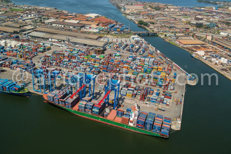 Africa photo — Vue aérienne d’un port maritime où un grand navire cargo chargé de conteneurs multicolores est amarré. Plusieurs grues géantes manipule