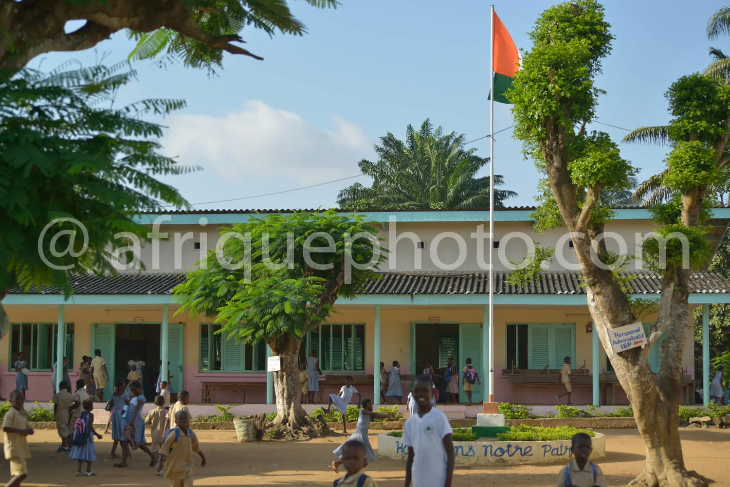 Africa photo — Une école primaire colorée avec des enfants en uniforme scolaire marchant dans une cour ombragée par des arbres. Un drapeau national fl