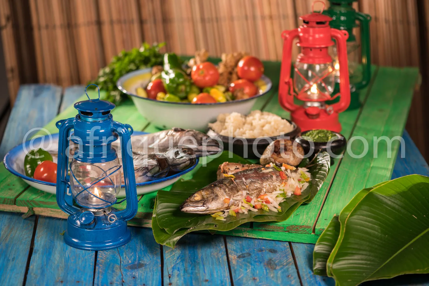Africa photo — Une table en bois bleu recouverte d’une nappe verte présente un poisson grillé enveloppé dans une feuille de bananier, accompagné de ri