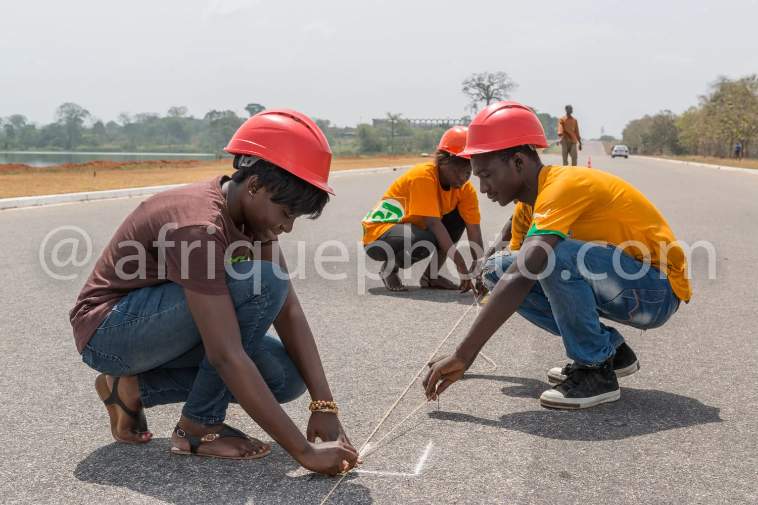 Africa photo — Trois personnes équipées de casques de sécurité rouges et de t-shirts orange s’activent sur une route en traçant des repères au sol ave