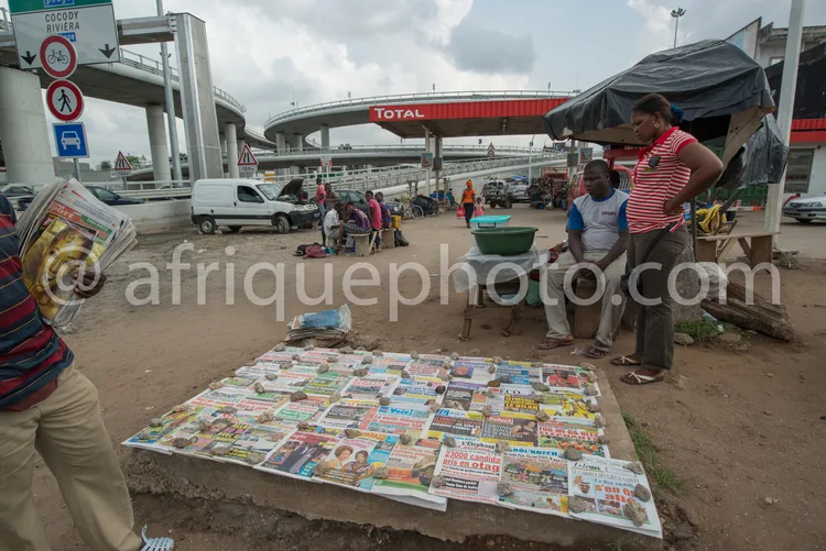 Africa photo — Une scène animée où deux personnes discutent près d’un étalage de journaux variés étalés sur le sol, avec une station-service Total en