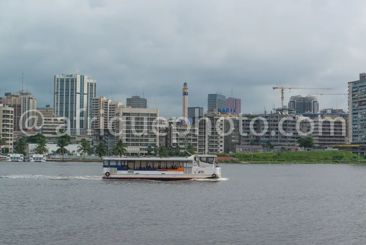 Africa photo — Un bateau de transport public, marqué STC, navigue sur un fleuve calme avec des passagers à bord, tandis qu'une skyline urbaine avec de