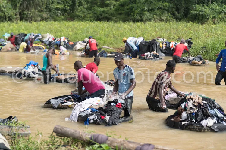 Africa photo — Quatre personnes assises dans une rivière peu profonde, entourées de sacs remplis de vêtements de différentes couleurs. Elles trient et